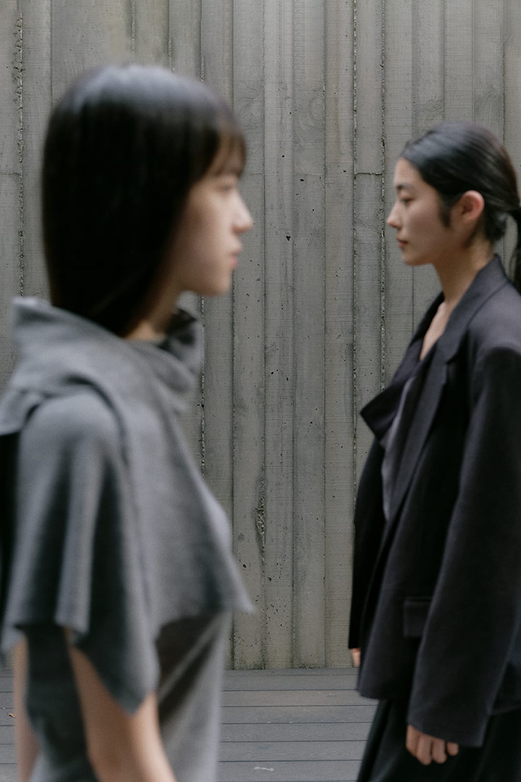 세로 결이 있는 콘크리트 건축 벽 앞을 그레이 드레이프 톱과 차콜 재킷 차림의 두 모델이 교차하는 모습