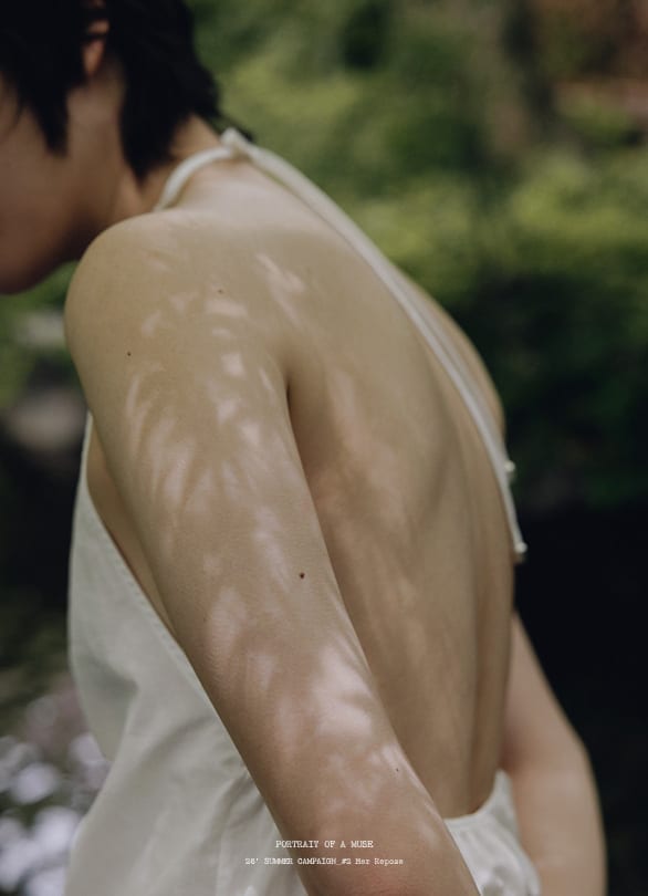 초록 나무와 물가를 배경으로 아이보리 홀터넥 오픈백 의상을 입은 모델의 등과 어깨